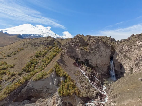 Dağ şelalesi ve Elbrus Dağı 'nın havadan görünüşü. Elbrus Dağı 'nın karlı, bulutsuz zirvesinin manzarası. Fırtınalı nehir dağın tepesinden vadiye akıyor ve yerden kırılıyor.