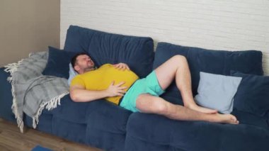 Sarı tişörtlü şişman adam evde yatağında uzanıyor ve hasta midesini okşuyor. Şişkinlik, aşırı yeme, kramp ve ağrı, ishal. Uygun beslenme