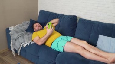 Şişman adam fiziksel egzersiz yapıyor. Kanepede uzanmak, akıllı telefondan video ile iletişim kurmak ve bisepsleri elle sallamak. Evde eğitim, karantina, salgın hastalık. Kilo kaybı, sağlıklı yaşam tarzı.