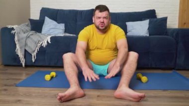 Sakallı tombul adam kilo vermek için fiziksel egzersiz yapıyor. Paspasa oturur ve ellerini ayak parmaklarına kadar uzatır. Evde eğitim, karantina, salgın hastalık. Kilo kaybı, sağlıklı yaşam tarzı.