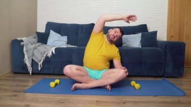 Sakallı tombul adam kilo vermek için fiziksel egzersiz yapıyor. Paspasın üzerine oturur ve kollarını farklı yönlere uzatır. Evde eğitim, karantina, salgın hastalık. Kilo kaybı, sağlıklı yaşam tarzı.