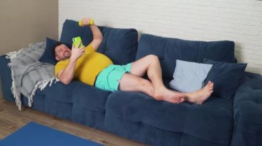 Şişman adam fiziksel egzersiz yapıyor. Kanepede uzanmak, akıllı telefondan video ile iletişim kurmak ve bisepsleri elle sallamak. Evde eğitim, karantina, salgın hastalık. Kilo kaybı, sağlıklı yaşam tarzı.