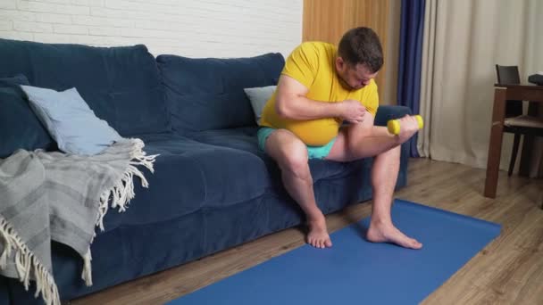 Pulchny mężczyzna z brodą robi ćwiczenia fizyczne dla utraty wagi. Siedzi na kanapie i macha rękami bicepsami. Trening w domu, kwarantanna, pandemia. Utrata masy ciała, zdrowy styl życia — Wideo stockowe