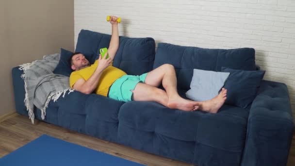 O homem gordo está a fazer exercício físico. Deitado no sofá, comunicando via vídeo no smartphone e sacudindo bíceps com a mão. Treino em casa, quarentena, pandemia. Perda de peso, estilo de vida saudável — Vídeo de Stock