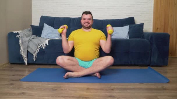Plump man is doing physical exercise for weight loss. Sits on sports mat and bends arms at the elbows with dumbbells in hands. Training at home, quarantine, pandemic. Weight loss, healthy lifestyle — Stock Video