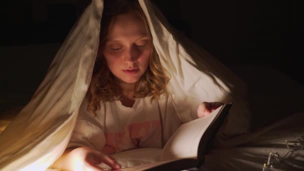 Vrouw leest boek onder deken in het donker en verlicht de pagina 's van het boek met zaklamp. Ze is verrast door wat ze heeft gelezen, glimlacht. Thuis lezen, sprookje, iets leren — Stockvideo