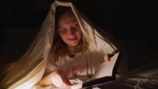 Frau liest Buch unter Decke in der Dunkelheit und beleuchtet die Seiten des Buches mit Taschenlampe. Sie ist überrascht von dem, was sie gelesen hat, lächelt. Lesen zu Hause, Märchen, etwas lernen — Stockvideo