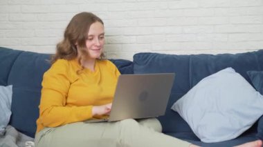 Kadın kanepede dizüstü bilgisayarıyla oturuyor ve bir şeyler yazıyor. Keyfi yerinde ve gülümsüyor. İyi haberler okuyor. İnternet 'te iletişim, e-öğrenme, evde çalışma, serbest çalışma, karantina, mesafe