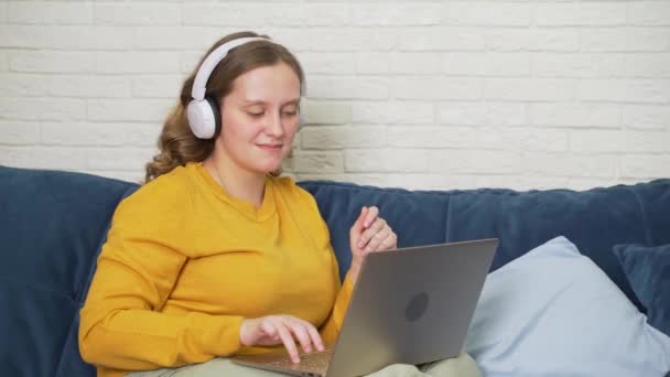 Mujer con auriculares blancos escucha música y baila. Ella está sentada en el portátil y trabajando, estudiando. Ella sonríe, sacude la cabeza y los tipos. Trabajo en laptop, negocios, estudio, cuarentena, distancia — Vídeos de Stock