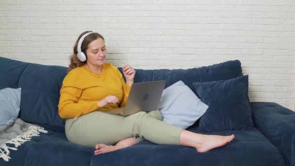 Kvinnan i vita hörlurar lyssnar på musik och danser. Hon sitter på laptop och jobbar och studerar. Hon ler, skakar huvud och typer. Arbete med bärbar dator, affärer, studier, karantän, distans — Stockvideo