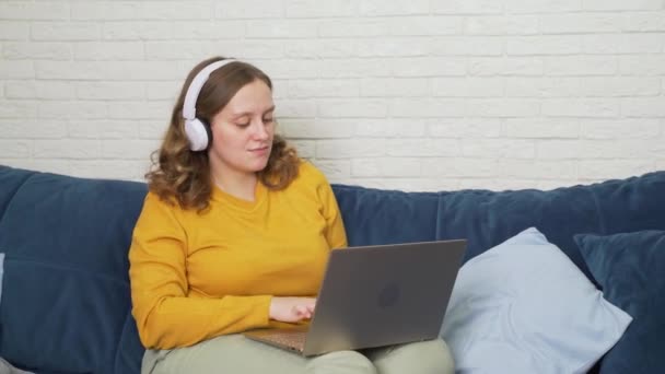 Woman in white headphones listens to music and dances. She is sitting at laptop and working, studying. She smiles, shakes head and types. Work on laptop, business, study, quarantine, distance — Stock Video