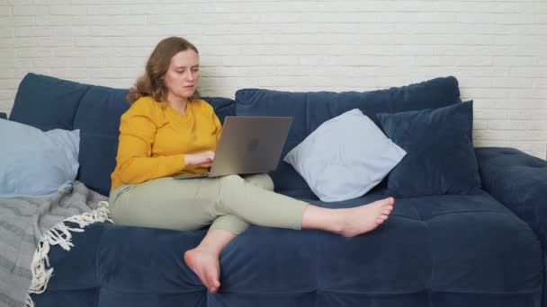 Mujer está sentada en el sofá con el ordenador portátil y escribir algo. Ella es seria e insatisfecha con algo. Agita la cabeza y extiende las manos. E-learning, trabajo en casa, freelance, cuarentena, distancia — Vídeos de Stock