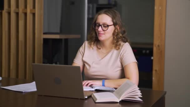 A szemüveges nő otthon ül az asztalnál és dolgozik, kommunikál kollégáival és barátaival videón keresztül az interneten keresztül egy laptopon. Otthoni munka, e-tanulás, karantén, távoktatás — Stock videók