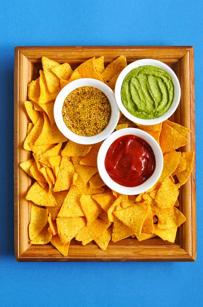 Heap of corn tortilla chips with french mustard, guacamole and ketchup served on a wooden board on a blue background. Top ivew.