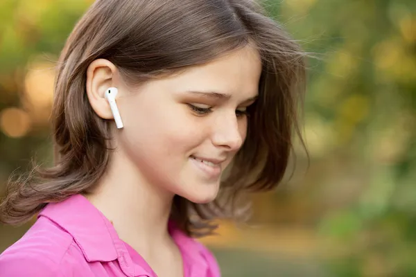Портрет прекрасной девушки в розовом, слушающей музыку через беспроводные наушники на открытом воздухе. Молодая девушка улыбается счастливой. Airpods. — стоковое фото