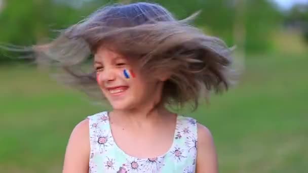 Barnglad flicka skakar gladeligen huvudet med sina kinder målade i flaggan av Frankrike, hennes hår flyger i olika riktningar. Långsam rörelse rolig skrattande liten blond liten flicka 6-8 år gammal utomhus — Stockvideo
