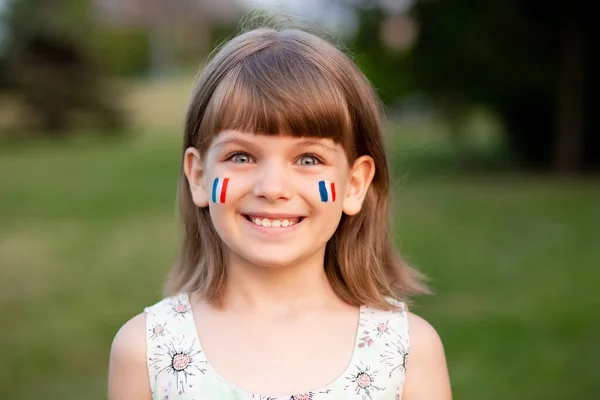 Retrato ao ar livre de pequena menina caucasiana com bochechas pintadas em cores bandeira da França e olhando para a câmera e sorrir. Jovem pintor. — Fotografia de Stock