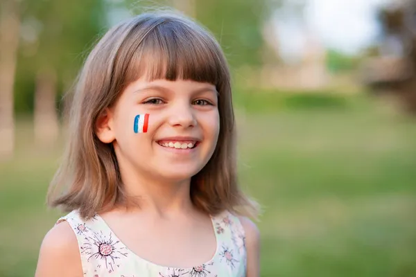 Ritratto all'aperto di bambina caucasica con le guance dipinte in Francia colori bandiera e guardando in macchina fotografica e sorriso. Giovane pittore. Foto Stock