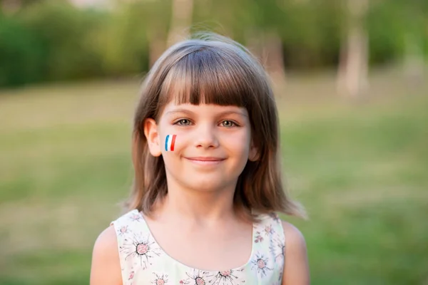Portrait extérieur de petite fille caucasienne aux joues peintes aux couleurs du drapeau français et regardant dans la caméra et le sourire. Jeune peintre. — Photo