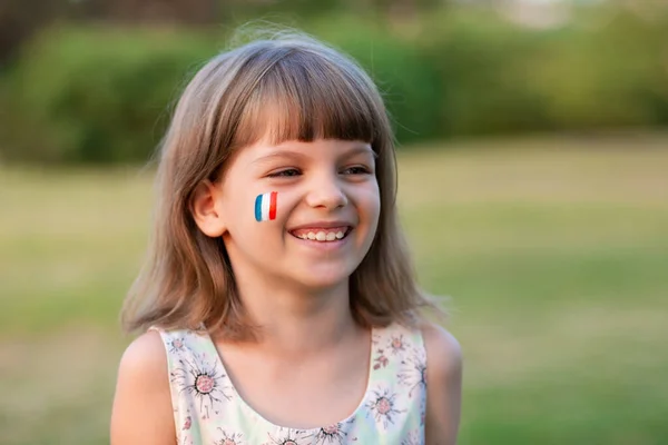 Utomhus porträtt av liten kaukasisk flicka med kinder målade i Frankrike flagga färger och tittar in i kameran och leende. Ung målare. — Stockfoto