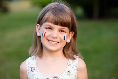 Fransız bayrağı rengine boyanmış yanaklı beyaz bir kızın açık hava portresi. Kameraya bakıp gülümsüyor. Genç ressam. Modern Sanat.