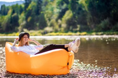 Nehir kıyısındaki sarı şişme kanepede uzanan güzel gülümseyen bir kızın yaz yaşam tarzı portresi. Rahatlamak, hava yatağında hayatın tadını çıkarmak. Tatil köyünde yatan tatilci kadın mutlu. Kız
