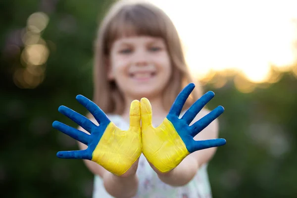 Joyful child girl waving hands painted in Ukraine flag colors and say hello outdoor at nature background, focus on hands