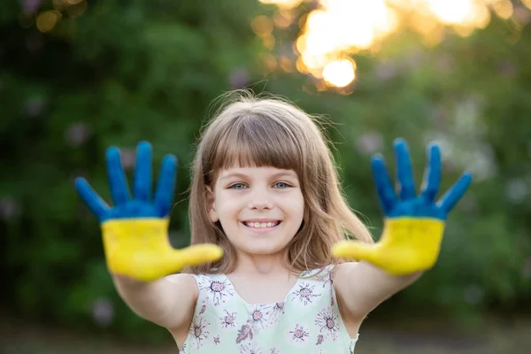 Joyful child girl waving hands painted in Ukraine flag colors and say hello outdoor at nature background, focus on hands