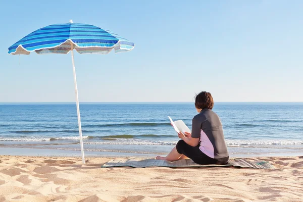 Frau mittleren Alters sitzt am Strand ein Buch zu lesen. unter einem b — Stockfoto