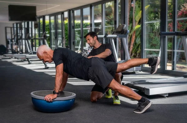 Un homme caucasien âgé, dans la salle de gym, avec un entraîneur de fitness personnel, en réadaptation, faire des exercices pour l'équilibre sur une balle de gymnastique. — Photo