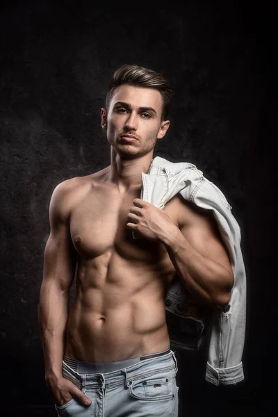 Europese atletische jongeman, poserend zonder bovenkleding, in sportvorm. Op een donkere textuur achtergrond. Jas op de schouder. — Stockfoto