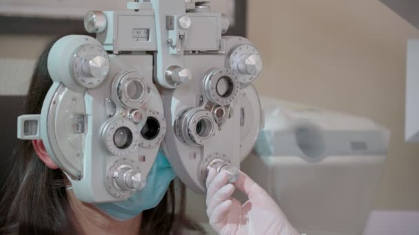 用光学光学仪器测量年轻妇女的视力 做视力测试的女人在大流行病中戴口罩的女孩和医生 — 图库视频影像