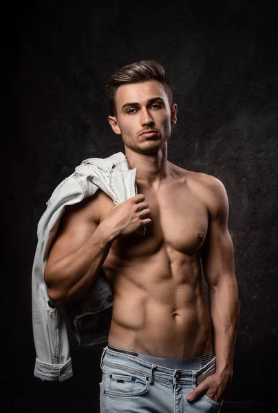 Europäischer athletischer junger Mann, ohne Oberbekleidung posierend, in sportlicher körperlicher Form. Auf dunklem, strukturiertem Hintergrund. Jacke auf der Schulter. — Stockfoto