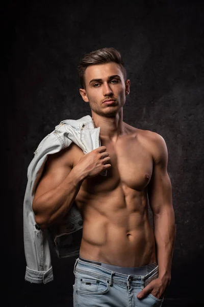 Europäischer athletischer junger Mann, ohne Oberbekleidung posierend, in sportlicher körperlicher Form. Auf dunklem, strukturiertem Hintergrund. Jacke auf der Schulter. — Stockfoto