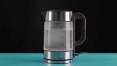 Modern cam elektrikli su ısıtıcısı, mavi bir masanın üzerinde, kaynaması için suyla dolu siyah bir arka plan. Onlara kaynar sudan içirilir..
