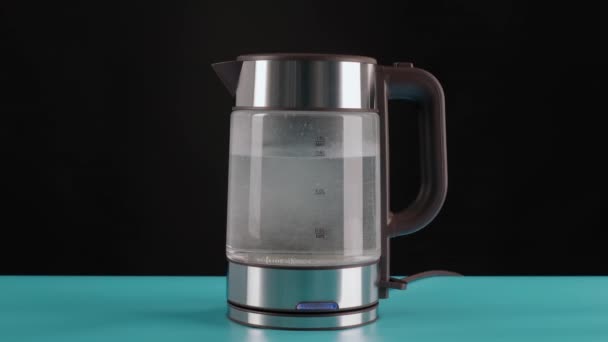 Un moderno hervidor eléctrico de vidrio, sobre una mesa azul, fondo negro, lleno de agua para hervir. Para hacer bebidas y agua hirviendo. — Vídeo de stock