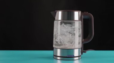Modern cam elektrikli su ısıtıcısı, mavi bir masanın üzerinde, kaynaması için suyla dolu siyah bir arka plan. Harekete geçti. Onlara kaynar sudan içirilir..