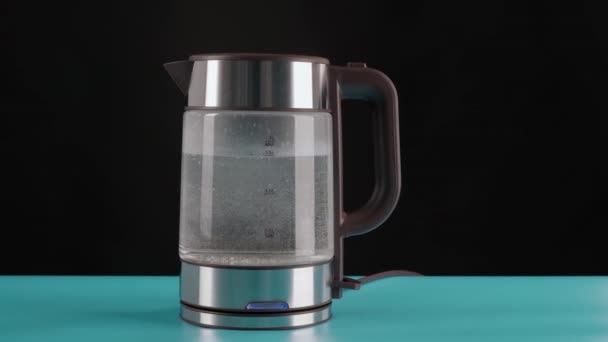Une bouilloire électrique en verre moderne, sur une table bleue, fond noir, remplie d'eau à ébullition. Pour faire des boissons et de l'eau bouillante, en mouvement. — Video