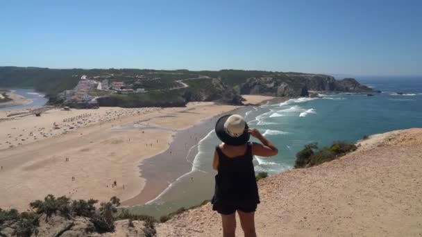 Vrouw met hoed van middelbare leeftijd staat op een berghelling en bewondert het prachtige uitzicht op het Portugese strand Odeceixe in de zomer met toeristen aan zee. — Stockvideo