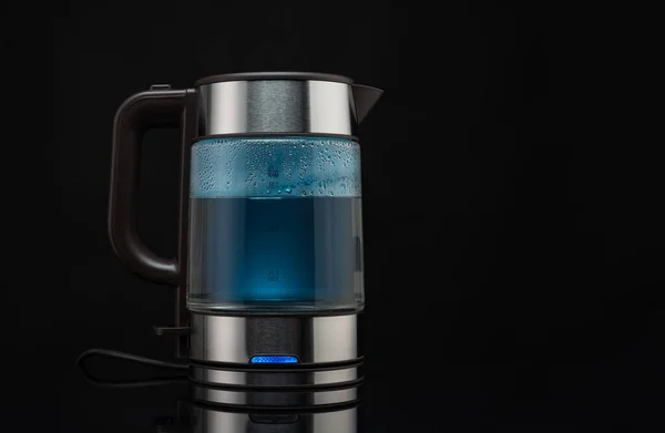 Electric glass kettle for boiling water for hot drinks on a black background with blue backlit water.