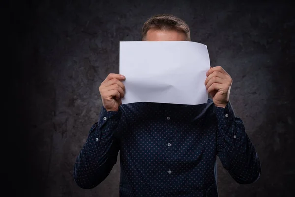 Un jeune Européen tient une feuille blanche vierge dans ses mains couvrant son visage. Pour la publicité sur un fond sombre — Photo