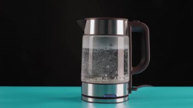 Modern cam elektrikli su ısıtıcısı, mavi bir masanın üzerinde, kaynaması için suyla dolu siyah bir arka plan. Onlara kaynar sudan içirilir. .