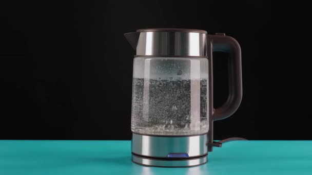 Une bouilloire électrique en verre moderne, sur une table bleue, fond noir, remplie d'eau à ébullition. Pour faire des boissons et de l'eau bouillante, en mouvement . — Video