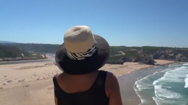 Orta yaşlı şapkalı bir kadın bir dağ yamacında duruyor ve yazın Portekiz sahilinin güzel manzarasına hayran kalıyor..