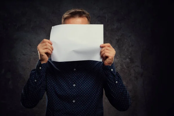 A young European guy holds a blank white sheet of paper in his hands covering his face. Copy space for advertising on a dark background — 图库照片