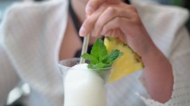 Tatilde olan orta yaşlı, beyaz bir kadın bir kamış ve bir dilim ananas aracılığıyla nane yapraklı hindistan cevizi kokteyli içiyor. Yakın plan..