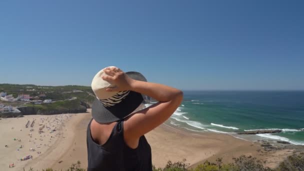 Vrouw met hoed van middelbare leeftijd staat op een berghelling en bewondert het prachtige uitzicht op het Portugese strand Odeceixe in de zomer met toeristen aan zee. — Stockvideo