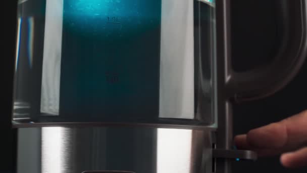 Пальцы человека нажимают на кнопку стеклянного чайника, чтобы вскипятить воду. Пузыри поднимаются в синем свете. низкое движение. — стоковое видео