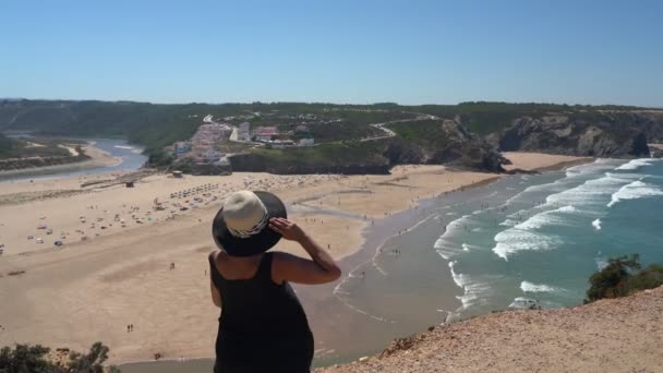 Vrouw met hoed van middelbare leeftijd staat op een berghelling en bewondert het prachtige uitzicht op het Portugese strand Odeceixe in de zomer met toeristen aan zee. — Stockvideo