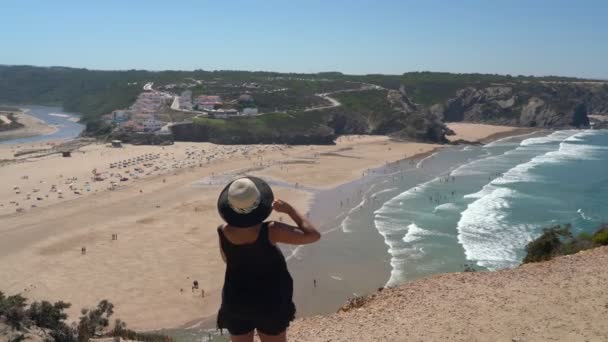 Nő egy középkorú kalap áll egy hegy lejtőjén, és csodálja a gyönyörű kilátás nyílik a portugál strand Odeceixe nyáron a turisták a tengernél. — Stock videók